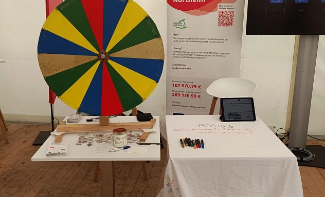 Infostand des Mobilen Demokratiezentrums beim Tag der Niedersachsen_Stand.jpg Informationsstand des Mobilen Demokratiezentrums im Landkreis Northeim in einem Veranstaltungszelt. Zu sehen sind ein buntes Glücksrad, Informationsmaterial, Buttons, eine Mitmachstation mit Tablet und Stiften sowie Roll-ups zum Projekt. Besucherinnen und Besucher konnten sich am Stand über Demokratiebildung informieren und an interaktiven Aktionen teilnehmen.