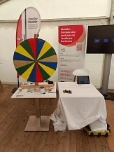 Informationsstand des Mobilen Demokratiezentrums im Landkreis Northeim in einem Veranstaltungszelt. Zu sehen sind ein buntes Glücksrad, Informationsmaterial, Buttons, eine Mitmachstation mit Tablet und Stiften sowie Roll-ups zum Projekt. Besucherinnen und Besucher konnten sich am Stand über Demokratiebildung informieren und an interaktiven Aktionen teilnehmen.&nbsp;&copy;&nbsp;Landkreis Northeim