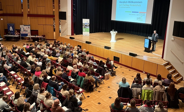 Kinderschutzfachtag 2025 1 Zu sehen ist die Stadthalle in Northeim, der Besucherraum ist mit Menschen gefüllt. Auf der Bühne steht Der Dezernent für Jugend und Soziales des Landkreises Northeim, Harald Rode. Auf der Präsentationsfolie im Hintergrund ist "Fachtag Kinderschutz" zu lesen.
