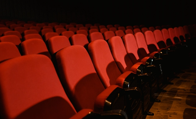 Reihe roter Kinosessel in einem dunklen Saal. Die Sitze sind leer und verlaufen perspektivisch nach hinten. Das gedimmte Licht lässt die Armlehnen und die Sitzreihen deutlich hervortreten, während der Hintergrund im Dunkel verschwindet.