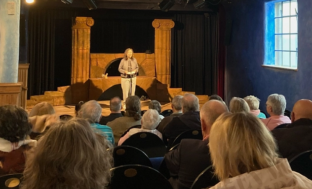 Landrätin begrüßt Ehrenamtliche Zu sehen ist eine Bühne in einem kleinen Theater, auf der die Landrätin steht und spricht. Das Foto ist von hinten im Saal aufgenommen, sodass man die Hinterköpfe von zahlreichen Personen sieht, die im Saal sitzen und zuhören.