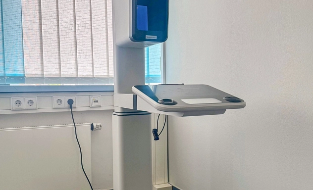 Lichtbildstation_Juili_2025 Eine moderne Lichtbildstation in einem Behördenraum. Das Gerät steht auf einem stabilen schwarzen Fuß und verfügt über einen Monitor, eine Kameraeinheit und eine integrierte Ablagefläche. Im Hintergrund befinden sich eine Fensterjalousie und mehrere Steckdosen an der Wand.