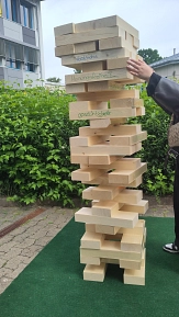 Ein übergroßer Jenga-Turm aus Holzklötzen steht im Freien auf einer grünen Matte. Eine Person zieht gerade vorsichtig einen Holzklotz aus dem wackeligen Turm. Auf mehreren Klötzen stehen Begriffe wie ‚Verständnis‘, ‚Meinungsfreiheit‘ und ‚Gerechtigkeit‘. Das Spiel symbolisiert, dass demokratische Werte das Fundament einer stabilen Gesellschaft bilden und jeder Baustein wichtig ist.&nbsp;&copy;&nbsp;Landkreis Northeim