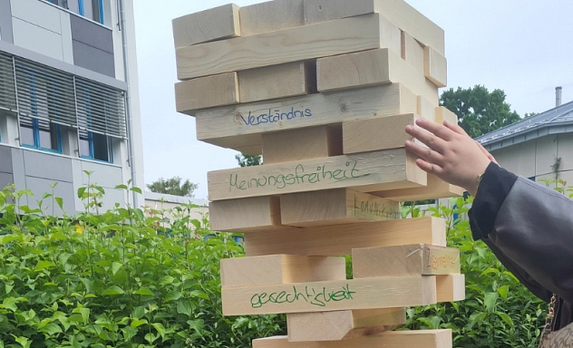 Mobiles Demokratiezentrum beim Aktionstag der BBS 1_ Jenga1_quer.png Ein übergroßer Jenga-Turm aus Holzklötzen steht im Freien auf einer grünen Matte. Eine Person zieht gerade vorsichtig einen Holzklotz aus dem wackeligen Turm. Auf mehreren Klötzen stehen Begriffe wie ‚Verständnis‘, ‚Meinungsfreiheit‘ und ‚Gerechtigkeit‘. Das Spiel symbolisiert, dass demokratische Werte das Fundament einer stabilen Gesellschaft bilden und jeder Baustein wichtig ist.