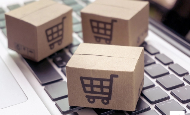 Drei kleine Pappkartons mit einem aufgedruckten Einkaufswagen-Symbol stehen auf einer Laptop-Tastatur. Die Kartons symbolisieren den Online-Handel und E-Commerce. Der Fokus liegt auf der Verbindung zwischen digitaler Bestellung und physischem Versand. In der unteren rechten Ecke befindet sich das Logo des Landkreises Northeim.