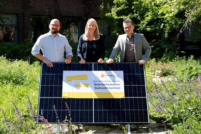 Drei Personen, Frithjof Look, Doreen Fragel und Jan-Christopher Linck (von links nach rechts), stehen in einem grünen Garten und halten gemeinsam ein großes Solarmodul. Auf dem Modul ist ein Plakat befestigt, auf dem steht: „Ist mein Dach für Solar geeignet? Jetzt herausfinden mit dem Solardachkataster Südniedersachsen.“ Im Hintergrund sind grüne Sträucher und ein Ziegelgebäude mit Fenstern zu sehen.&nbsp;&copy;&nbsp;Energieagentur Göttingen e.V.