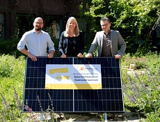 Drei Personen, Frithjof Look, Doreen Fragel und Jan-Christopher Linck (von links nach rechts), stehen in einem grünen Garten und halten gemeinsam ein großes Solarmodul. Auf dem Modul ist ein Plakat befestigt, auf dem steht: „Ist mein Dach für Solar geeignet? Jetzt herausfinden mit dem Solardachkataster Südniedersachsen.“ Im Hintergrund sind grüne Sträucher und ein Ziegelgebäude mit Fenstern zu sehen.