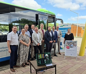 Zehn Personen – eine Frau und neun Männer – stehen vor einem Linienbus des Verkehrsverbunds Süd-Niedersachsen (ZVSN) und posieren für ein Gruppenfoto. Vor ihnen ist ein automatisches Fahrgastzählsystem mit Kamera- und Sensortechnik auf einem Tisch aufgebaut. Im Hintergrund stehen ein ZVSN-Roll-up sowie ein Informationsaufsteller zum Projekt. Die Aufnahme entstand bei der Vorstellung der neuen Fahrgastzählsysteme für den öffentlichen Nahverkehr.&nbsp;&copy;&nbsp;Landkreis Northeim