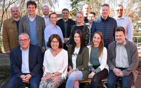 Eine Gruppe von 14 Personen posiert gemeinsam für ein Foto im Freien. Die Personen sind in zwei Reihen angeordnet: fünf Personen sitzen auf einer Bank in der vorderen Reihe, während neun weitere dahinterstehen. Alle blicken in die Kamera und lächeln. Die Kleidung reicht von formellen Sakkos bis hin zu legeren Jacken und Pullovern. Im Hintergrund sind Bäume, ein Geländer und einige Gebäude mit roten Dächern zu sehen, was auf eine ländliche oder kleinstädtische Umgebung hindeutet. Die Atmosphäre wirkt freundlich und entspannt.&nbsp;&copy;&nbsp;Landkreis Northeim