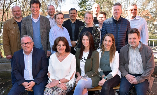 Runder Tisch Radverkehr Eine Gruppe von 14 Personen posiert gemeinsam für ein Foto im Freien. Die Personen sind in zwei Reihen angeordnet: fünf Personen sitzen auf einer Bank in der vorderen Reihe, während neun weitere dahinterstehen. Alle blicken in die Kamera und lächeln. Die Kleidung reicht von formellen Sakkos bis hin zu legeren Jacken und Pullovern. Im Hintergrund sind Bäume, ein Geländer und einige Gebäude mit roten Dächern zu sehen, was auf eine ländliche oder kleinstädtische Umgebung hindeutet. Die Atmosphäre wirkt freundlich und entspannt.