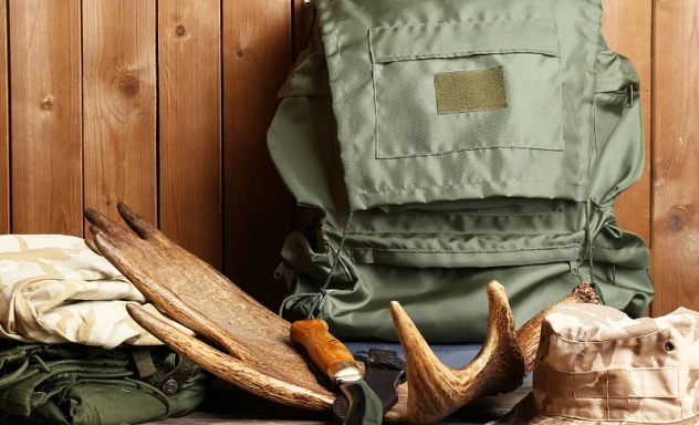 Zu sehen ist Jagdausstattung: Ein olivfarbener Rucksack, der an eine Holzwand lehnt im Hintergrund. Davor liegt ein Messer, ein Tarnhut und ein Geweih.