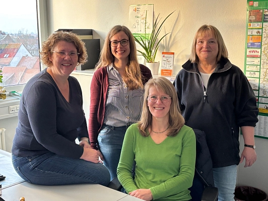 Team des Senioren- und Pflegestützpunktes 2025 Gruppenfoto von vier lächelnden Frauen in einem Büro. Eine Frau sitzt vorne auf einem Stuhl, die anderen stehen bzw. sitzen dahinter. Im Hintergrund sind Fenster mit Blick auf Dächer, eine Pflanze, Flyer und ein Info-Poster an der Wand zu sehen. Alle wirken freundlich und einladend. © Landkreis Northeim