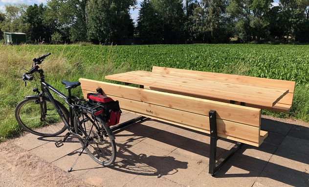 Tisch und Bänke bei Holtensen Vor dem Hintergrund einer Wiese und Bäumen steht eine Tisch-Bank-Kombination aus Holz. Davor steht ein Fahrrad.