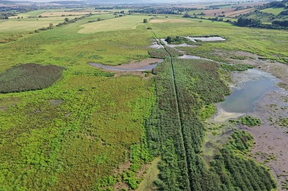 Luftaufnahme des EU-Vogelschutzgebiets Leinetal bei Salzderhelden. Die Landschaft besteht aus einer weitläufigen, teils sumpfigen Wiesenlandschaft mit kleinen Wasserflächen und dicht bewachsenen Schilfzonen. Ein schmaler, von Vegetation gesäumter Wassergraben verläuft durch das Gebiet. Im Hintergrund erstrecken sich landwirtschaftlich genutzte Felder und sanfte Hügel. Die verschiedenen Feuchtgebiete und offenen Flächen bieten wertvollen Lebensraum für zahlreiche Vogelarten.&nbsp;&copy;&nbsp;Landkreis Northeim