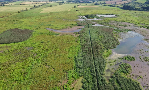 Vogelschutzgebiet "Leinetal bei Salzderhelden" Luftaufnahme des EU-Vogelschutzgebiets Leinetal bei Salzderhelden. Die Landschaft besteht aus einer weitläufigen, teils sumpfigen Wiesenlandschaft mit kleinen Wasserflächen und dicht bewachsenen Schilfzonen. Ein schmaler, von Vegetation gesäumter Wassergraben verläuft durch das Gebiet. Im Hintergrund erstrecken sich landwirtschaftlich genutzte Felder und sanfte Hügel. Die verschiedenen Feuchtgebiete und offenen Flächen bieten wertvollen Lebensraum für zahlreiche Vogelarten.