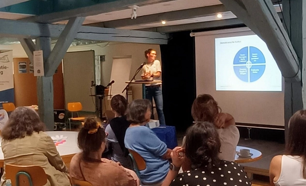 Vortrag Demokratische Kultur Eine Person hält in einem Veranstaltungsraum einen Vortrag vor Publikum. Im Hintergrund ist eine Präsentationsfolie auf einer Leinwand zu sehen; die Teilnehmenden sitzen an Tischen und hören zu.