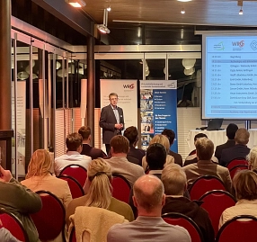 Vortrag Technologieberatung Das Bild zeigt eine Veranstaltung in einem Konferenzraum. Im Vordergrund sitzt ein Publikum auf roten Stühlen, während im Hintergrund ein Redner vor einem großen Bildschirm steht. Auf dem Bildschirm ist eine Agenda der Veranstaltung erkennbar, die unter anderem Begrüßung und Vorträge verschiedener Unternehmen enthält. Im Hintergrund sind Banner der Wirtschaftsförderung und eines weiteren Veranstalters zu sehen. Die Szene ist beleuchtet und vermittelt eine professionelle Atmosphäre. © Landkreis Northeim