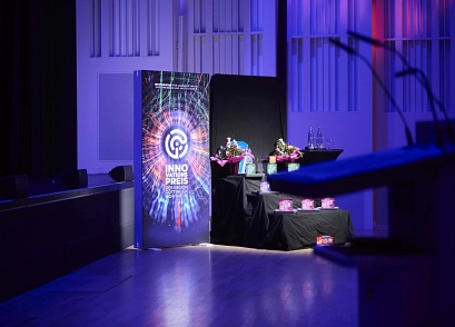 Bühne bei der Verleihung des Innovationspreises der Region Göttingen Northeim: Ein beleuchteter Aufsteller steht neben einem Tisch mit Blumensträußen, Glaspreisen und Wasserflaschen. Die Szene ist in blau-violettes Licht getaucht.&nbsp;&copy;&nbsp;Marco Bühl