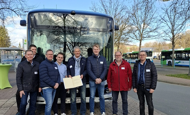 ZVSN Elektrobusse im Alltagstest Vertreter von Landkreis und Verkehrsbetrieben stehen vor einem Linienbus und präsentieren eine Urkunde bei einem Termin am Busbahnhof.