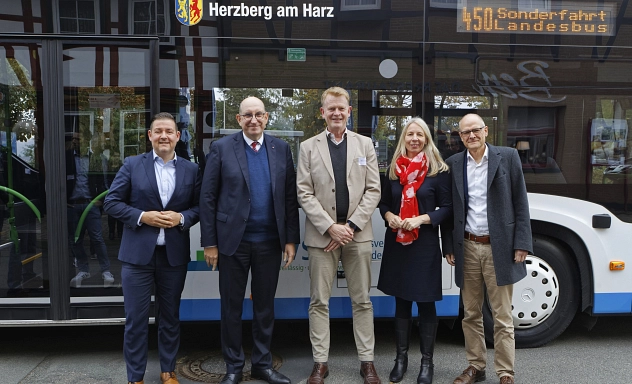 ZVSN: Neue Direktverbindung Harz - Braunlage Gruppenfoto Fünf Personen, vier Männer und eine Frau, stehen vor einem weißen Linienbus mit der Aufschrift ‚Herzberg am Harz‘ und der Linienanzeige ‚450 Sonderfahrt Landesbus‘. Die Gruppe besteht aus drei Männern in Anzügen, einer Frau mit rotem Schal und einem weiteren Mann im Mantel. Sie lächeln in die Kamera, der Bus dient als Hintergrund.